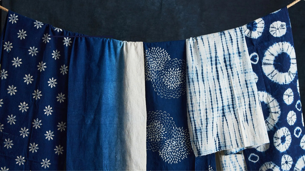 A close-up of several indigo-dyed fabric pieces hanging side by side, showing different patterns including white floral prints, tie-dye stripes, and circular motifs on a dark blue background.