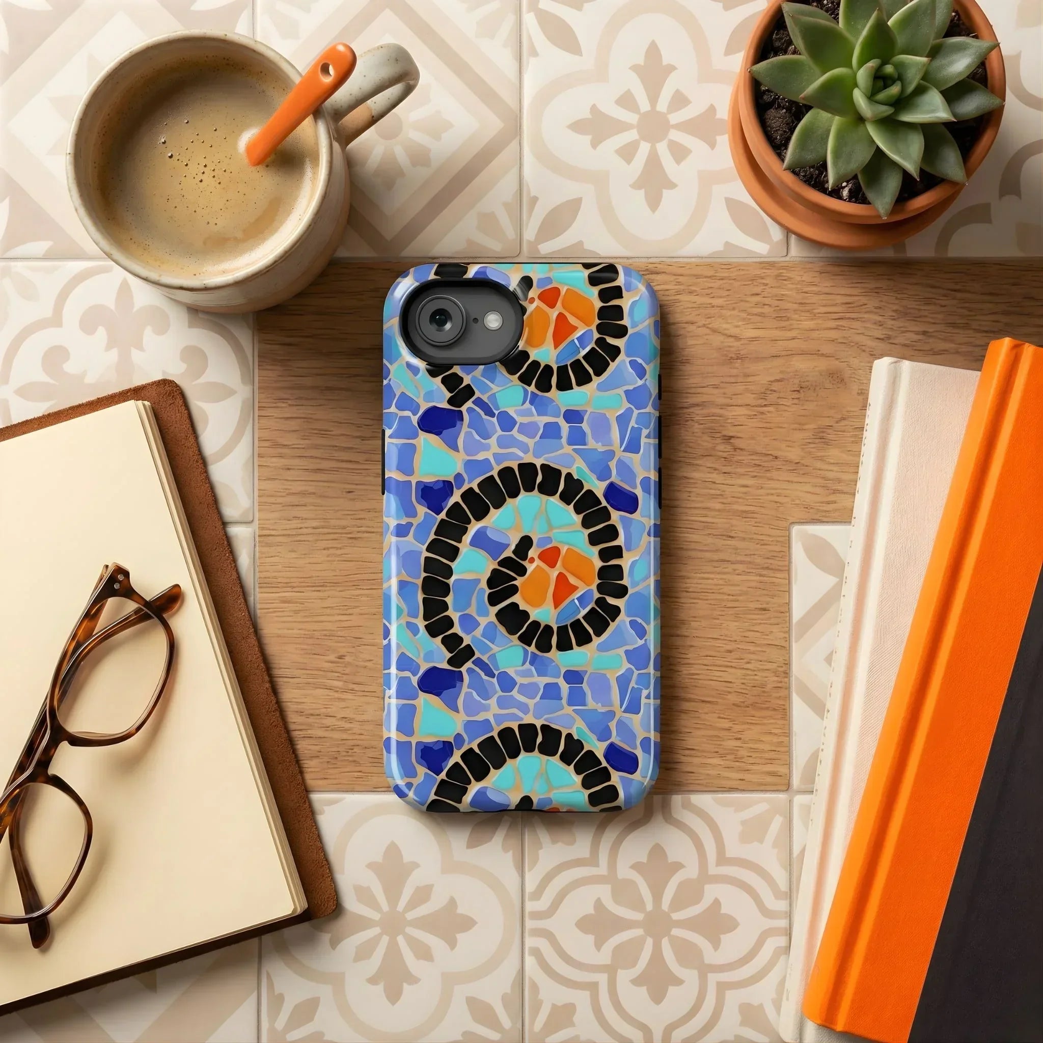 Phone case with Spanish mosaic design on a desk with coffee, glasses, and books.