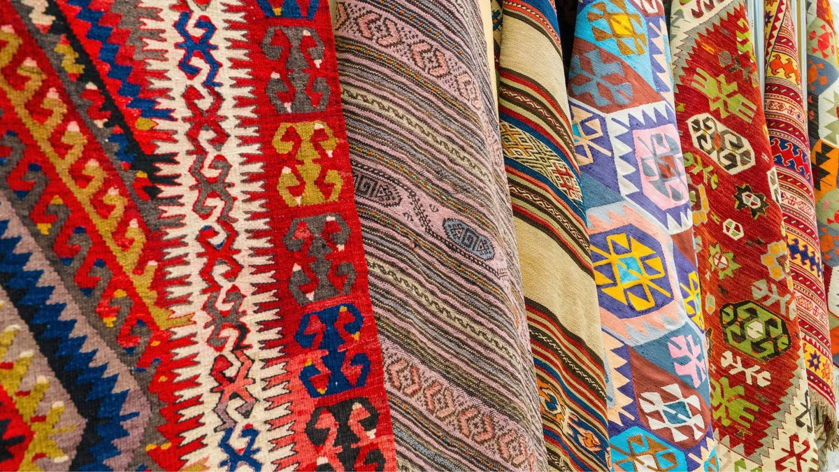 A close-up view of multiple hanging kilim rugs with intricate geometric and tribal patterns in red, blue, beige, brown, and white.