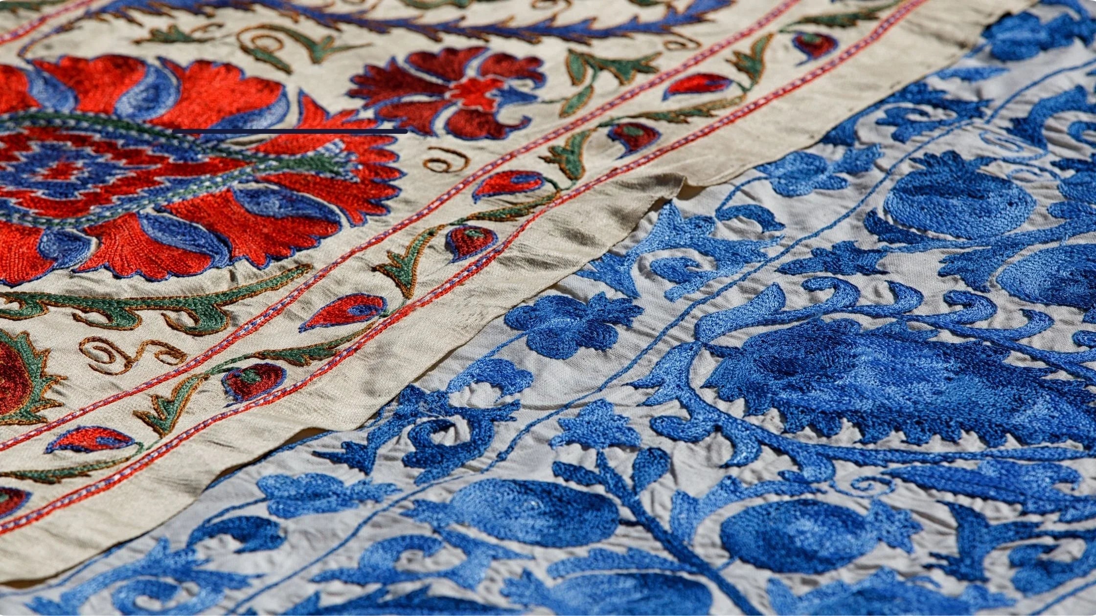 Close-up of an embroidered textile with cream fabric, red and blue floral motifs, and blue ornamental embroidery across a border.