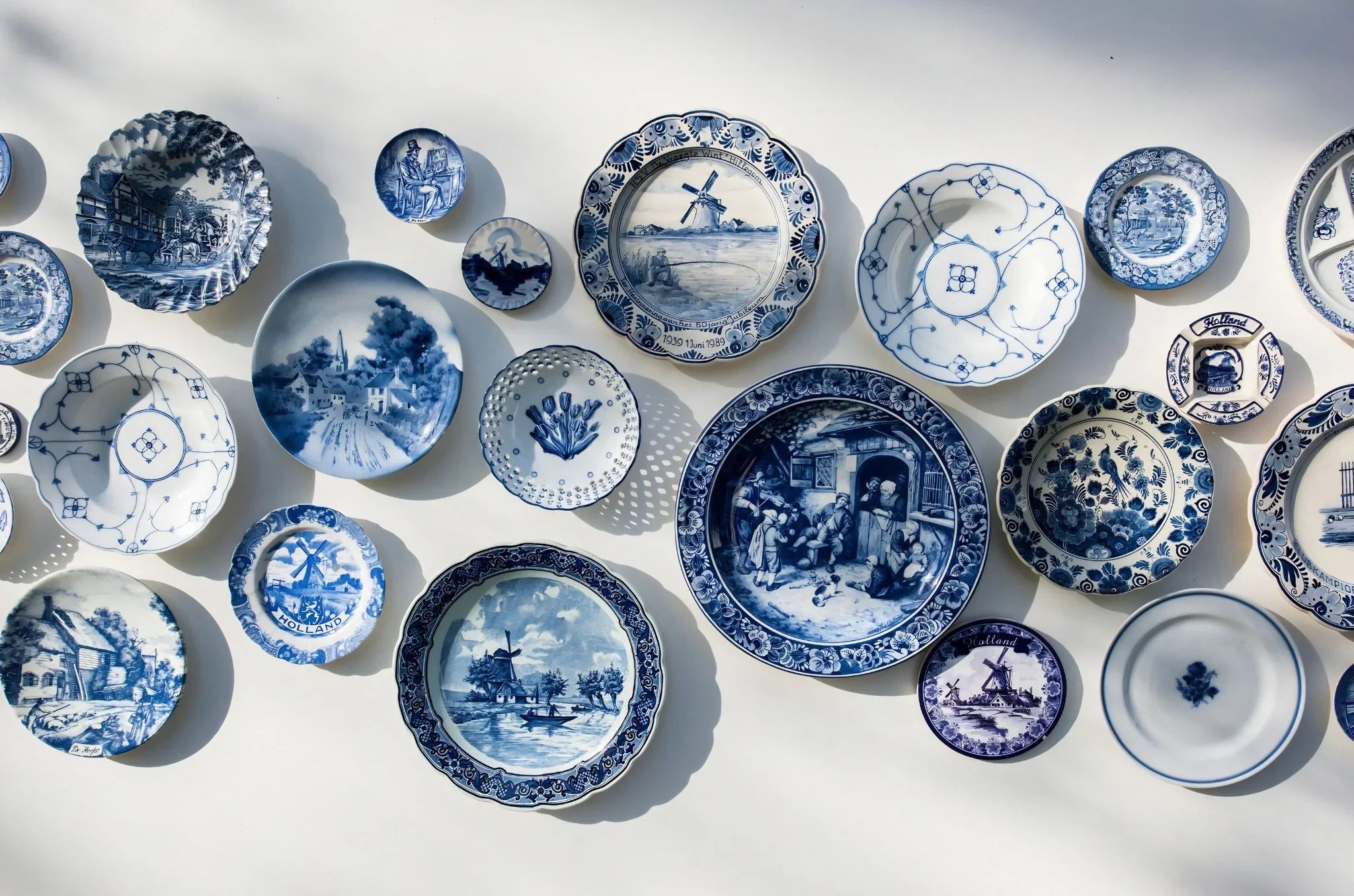 An assortment of blue and white porcelain plates and small dishes with various hand-painted traditional Dutch and European landscape designs, arranged on a white wall.
